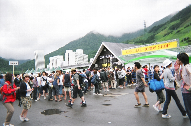 フジロックフェスティバルの公式グッズ売り場。アーティストのグッズ売り場ではありません。左に「列最後尾」のプラカードが見えます。こういうものが事前に用意されるくらいに混雑します。1時間待ちも珍しくありません。デザインのいいTシャツや女性用サイズのTシャツなどはすぐ売り切れます。デザインは何十種類もあります。通常はテントの上のサンプルのTシャツを何十着も掲げてあります。このときは雨が降っていたので掲げられていません。