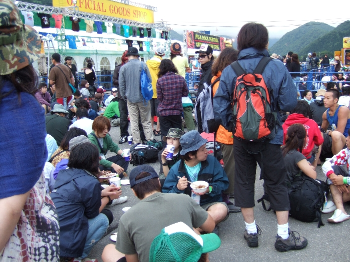 場外ショップエリアで昼食を取りながらオフィシャル・グッズ売り場に並ぶ人。
