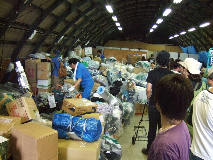 リストバンド交換所の近くにある宅配便受付の中。荷物が山積みにされており、ヤマト運輸や西武運輸の社員、受取人（フジ・ロック参加者）が荷物を探しています。