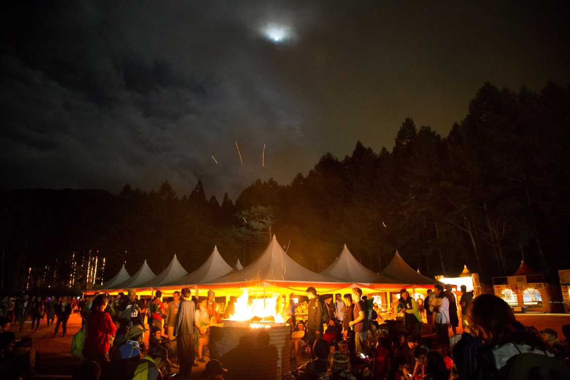 上に月、左にボードウォークの電飾、中央にたき火。