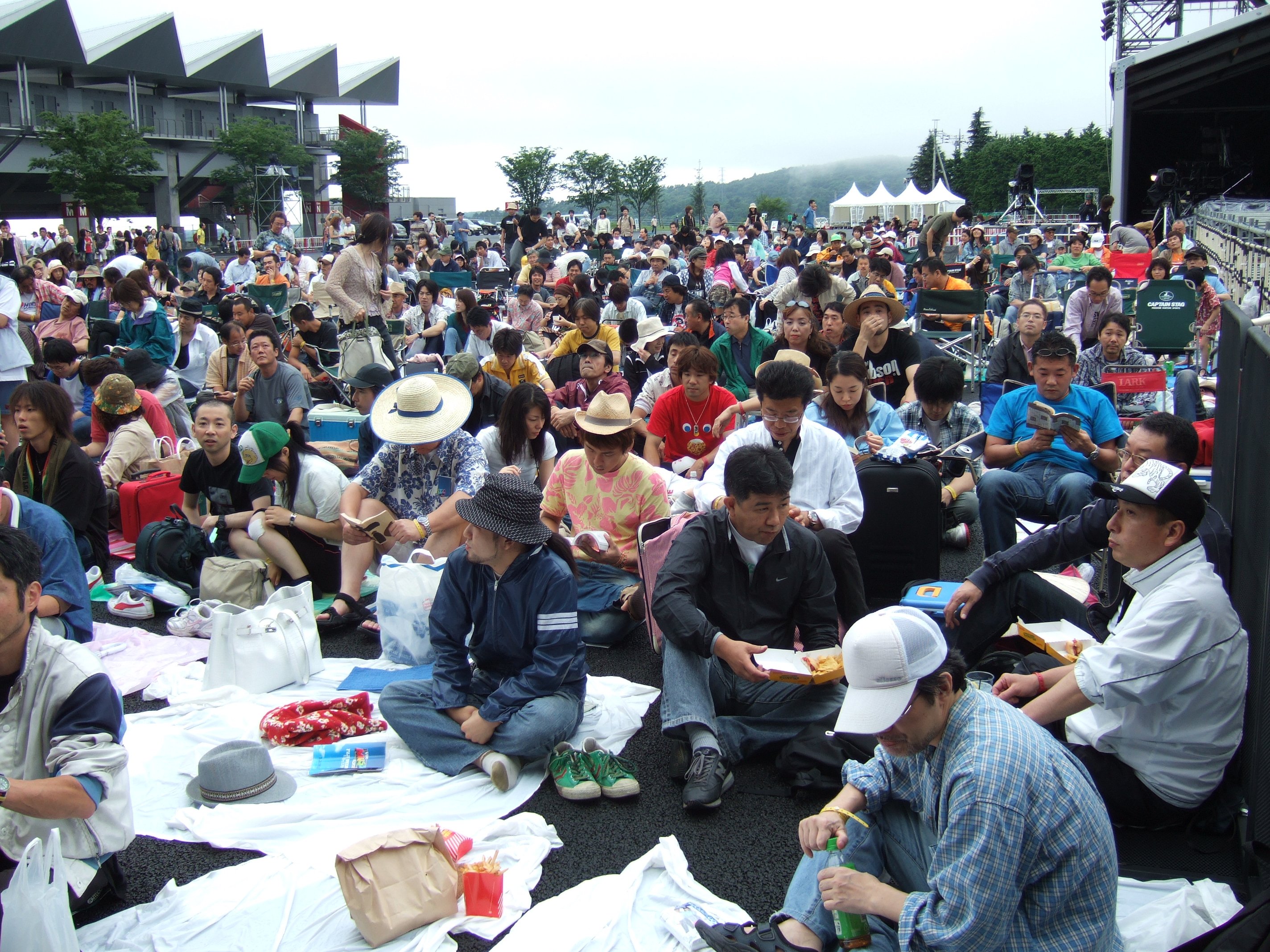 最前列近くから客席を撮影。1日目午前10時ごろ。まだ最初のアーティストが始まっていない時間です。このときは、客席に関するルールは何もない状態でした。最前列でもレジャーシートを敷き、いすを置いて座っている人がほとんどでした。この後、後方から若い観客がすき間を埋めるように前に進んできます。そこで、当初から前の方にいた40代、50代の人が「何時から並んだと思ってんだよ」と文句を言うと、フェスティバル慣れした若い観客が「指定席じゃないでしょう」と対抗していました。40代、50代の人にとっては、せっかく朝早く並んで確保した前の列を、あとから割り込んでくる若者に奪われるのが我慢ならなかったのでしょう。しかし、40代、50代の人にとっては、自分たちが70年代に体験してきたコンサートの「慣習」に従っているだけです。早く来た人がいい席を確保する、あとから来た人は後ろの席で我慢する、というのは、それ自体としては妥当なルールで、「あとから来た若いヤツは後ろで見てろ」と言うのは非常識な物言いとは言えません。しかし、フジロックフェスティバルやサマーソニックに慣れた20代の若者は、自分が見たいアーティストが来れば前に行き、それほどでもないアーティストではあっさり後ろに下がるというのが「慣習」になっています。最初から最後まで前の列に居続けることはありません。ウドー・ミュージック・フェスティバルは中高年をターゲットにしたロック・フェスティバルであるため、世代間の認識の違いがどこかで現れ、それがいさかいの原因になるのはやむを得ないところがありました。この件では、しばらく時間をおいてから主催者側の人が現れ「ステージ前はスタンディング形式とする」「レジャーシート、いすによる場所の占拠は不可」とのルール設定が宣言されました。若い人を中心に拍手が起き、後方から次々と人が押し寄せました。当初からいた人は押し出されるように態勢を縮小していきましたが、このとき若い人から「さっさとどけよ」という声がありました。こうした挑発的な態度が無用なトラブルを生むことを、若い人は自覚するべきでしょう。いくらそれまでの行為に落ち度がなくても。これによって、不快な印象が結局若者の方に残ってしまいます。